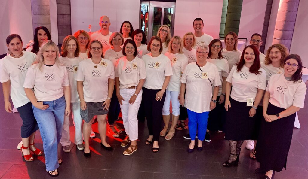 Gruppe von Personen in weißen T-Shirts mit schwarzem Logo von der Rheinhessen Sparkasse, stehend in einem Raum mit rosa und grüner Beleuchtung.