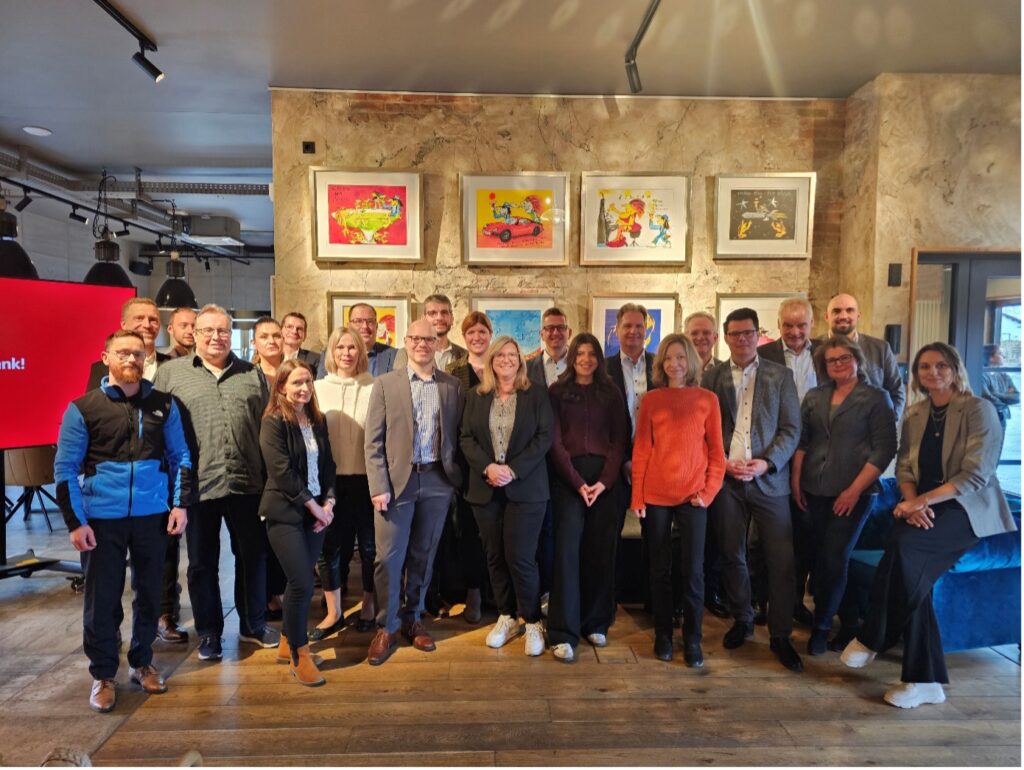 Gruppe von Personen steht in einem Raum mit Holzfußboden vor einer Wand mit vier farbigen, gerahmten Bildern.