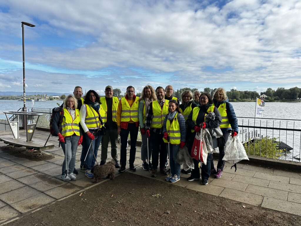 Gruppe von Personen mit gelben Warnwesten steht auf gepflastertem Weg am Flussufer bei bewölktem Himmel.